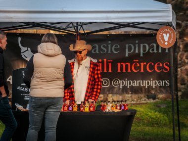 Kovboy şapkası ve ekose gömlekli sakallı bir satıcı açık hava pazarında renkli acı soslarla dolu bir masanın arkasında duruyor. Poster ve etkileşimli müşteriler görünür..