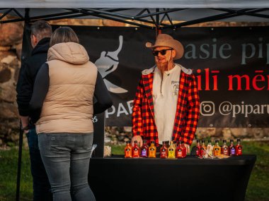Kovboy şapkası ve ekose gömlekli sakallı bir satıcı açık hava pazarında renkli acı soslarla dolu bir masanın arkasında duruyor. Poster ve etkileşimli müşteriler görünür..