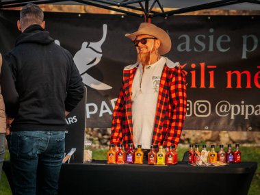 Kovboy şapkası ve ekose gömlekli sakallı bir satıcı açık hava pazarında renkli acı soslarla dolu bir masanın arkasında duruyor. Poster ve etkileşimli müşteriler görünür..