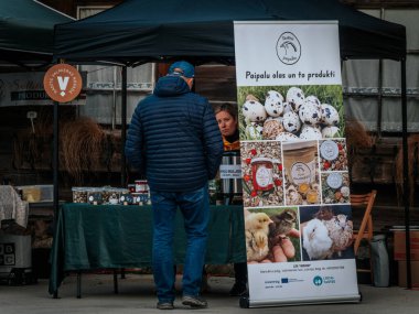 Bir satıcı yerel bir açık hava pazarında müşterilere yiyecek kavanozları sunar. Çocuklu ve bebek arabalı aileler ahşap bir standın etrafında toplanıyorlar. Konforlu ve toplum odaklı. 17.05.2025 Valmiera, Letonya