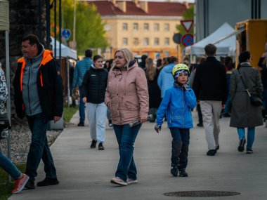 Yetişkinler ve çocuklar Valmiera Müzesi Gece Pazarı Meydanı 'nda dolanıyor. Mavi ceketli ve kasklı bir çocuk ön planda göze çarpıyor. Beyaz çadırlar. 17.05.2025 Valmiera, Letonya