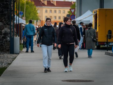 Kalabalık, kırmızı kiremitli çatısı olan bir binanın yakınındaki açık hava pazarında geziniyor. Kasklı bisikletler yakınlarda park halinde ve Letonya 'da 17.05.2025 Valmiera yolu boyunca satıcıların çadırları kurulmuş durumda.
