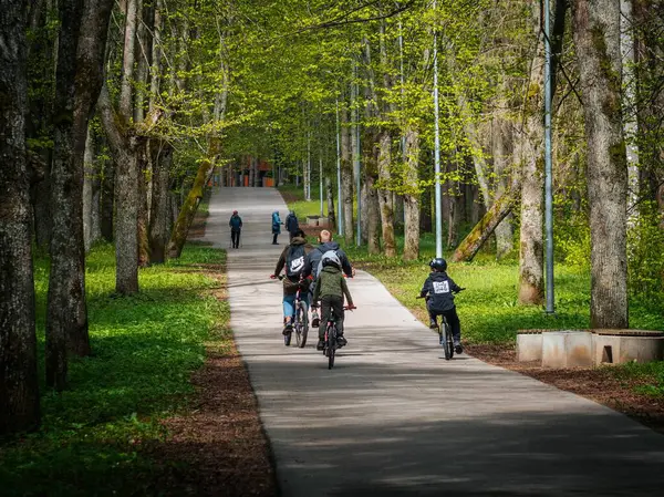 Aralarında çocuklar ve yetişkinlerin de yer aldığı bir grup bisikletçi, uzun ağaçlar ve taze bahar yapraklarıyla çevrili asfalt bir orman yolu boyunca ilerliyorlar. İnsanlar önde yürür..