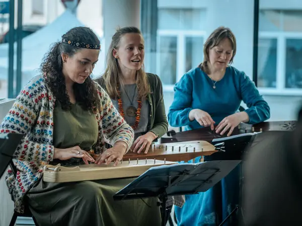 Geleneksel giysiler içinde bir grup kadın, kültürel bir mekanda ahşap ip enstrümanlarıyla gösteri yapıyorlar. Sayfa notaları ve işaretler resmi ya da tarihi bir olayı gösterir.