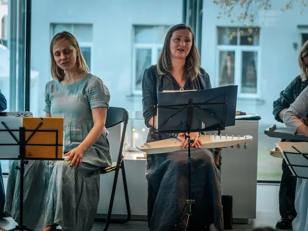 Geleneksel giysiler içinde bir grup kadın, kültürel bir mekanda ahşap ip enstrümanlarıyla gösteri yapıyorlar. Sayfa notaları ve işaretler resmi ya da tarihi bir olayı gösterir.