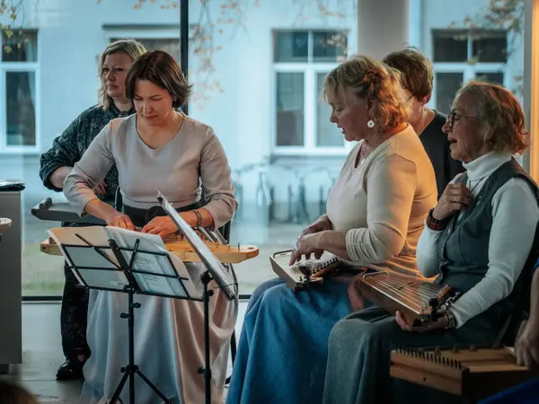 Geleneksel giysiler içinde bir grup kadın, kültürel bir mekanda ahşap ip enstrümanlarıyla gösteri yapıyorlar. Sayfa notaları ve işaretler resmi ya da tarihi bir olayı gösterir.