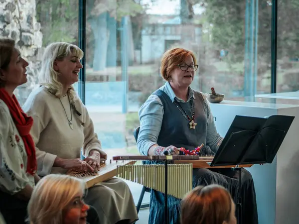 Geleneksel giysiler içinde bir grup kadın, kültürel bir mekanda ahşap ip enstrümanlarıyla gösteri yapıyorlar. Sayfa notaları ve işaretler resmi ya da tarihi bir olayı gösterir.