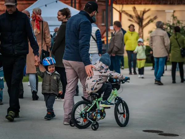 Bir çocuk, diğerleri Valmiera Müzesi Gece Parkı 'nda anne ve babasıyla açık hava ortamında dolaşırken küçük yeşil bir bisiklete biniyor. İnsanlar toplanır.