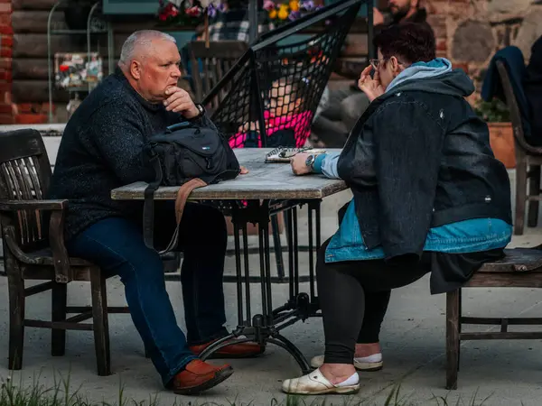 İnsanlar açık bir kafede kırsal ahşap masalarda oturur, bazıları kahve içerken diğerleri pizza yer. Ön plandaki sahne dalgındır..