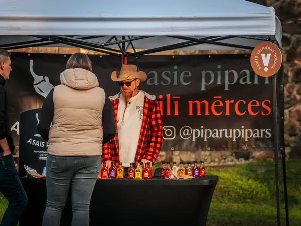 Kovboy şapkası ve ekose gömlekli sakallı bir satıcı açık hava pazarında renkli acı soslarla dolu bir masanın arkasında duruyor. Poster ve etkileşimli müşteriler görünür..