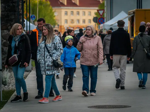 Yetişkinler ve çocuklar Valmiera Müzesi Gece Pazarı Meydanı 'nda dolanıyor. Mavi ceketli ve kasklı bir çocuk ön planda göze çarpıyor. Beyaz çadırlar. 