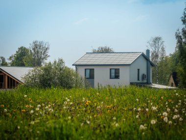 Karahindiba tohumu başları ve sarı çiçeklerle dolu parlak yeşil bir çayır biri hala yapım aşamasında olan iki modern eve doğru uzanır, açık mavi gökyüzüne karşı..