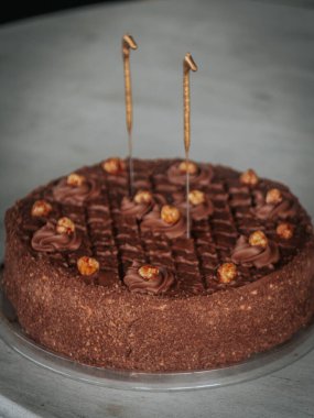  Yuvarlak çikolatalı pasta kıvrımlı ve karamelize edilmiş fındıklı. Gri bir masanın üstünde iki aydınlatılmamış maytap var. Bayramlık bir olay veya doğum günü için ideal..