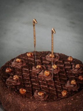  Yuvarlak çikolatalı pasta kıvrımlı ve karamelize edilmiş fındıklı. Gri bir masanın üstünde iki aydınlatılmamış maytap var. Bayramlık bir olay veya doğum günü için ideal..