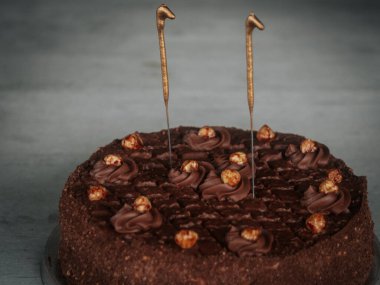  Yuvarlak çikolatalı pasta kıvrımlı ve karamelize edilmiş fındıklı. Gri bir masanın üstünde iki aydınlatılmamış maytap var. Bayramlık bir olay veya doğum günü için ideal..