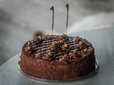 Yuvarlak çikolatalı pasta kıvrımlı ve karamelize edilmiş fındıklı. Gri bir masanın üstünde iki aydınlatılmamış maytap var. Bayramlık bir olay veya doğum günü için ideal..
