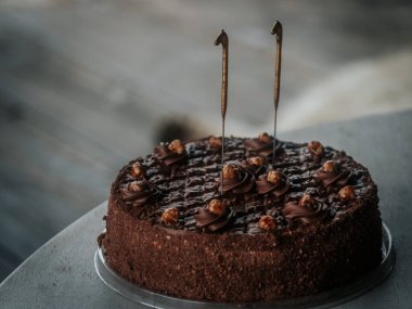  Yuvarlak çikolatalı pasta kıvrımlı ve karamelize edilmiş fındıklı. Gri bir masanın üstünde iki aydınlatılmamış maytap var. Bayramlık bir olay veya doğum günü için ideal..