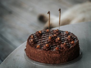  Yuvarlak çikolatalı pasta kıvrımlı ve karamelize edilmiş fındıklı. Gri bir masanın üstünde iki aydınlatılmamış maytap var. Bayramlık bir olay veya doğum günü için ideal..