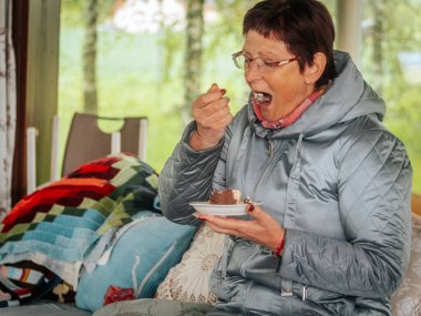 Açık gri örtülü ceketli ve gözlüklü bir kadın kapalı bir bankta oturuyor, elinde çikolatalı pasta tabağı, yastıklarla ve yumuşak ışıklandırmalarla.