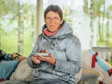 Açık gri örtülü ceketli ve gözlüklü bir kadın kapalı bir bankta oturuyor, elinde çikolatalı pasta tabağı, yastıklarla ve yumuşak ışıklandırmalarla.