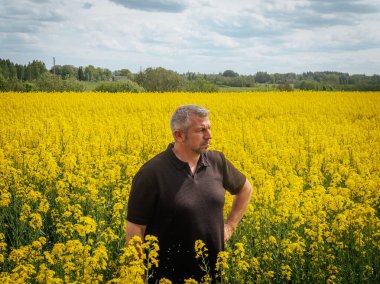 Koyu renk gömlekli düşünceli bir adam bulutlu bir bahar gökyüzünün altında çiçek açan sarı çiçeklerle çevrili geniş bir kolza tohumu tarlasında duruyor..