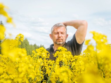  Düşünceli bir ifadeye sahip bir adam çiçek açan bir kolza tohumu tarlasında duruyor, bir eli başının arkasında, yumuşak, parçalı bulutlu bir gökyüzünün altında dinleniyor..