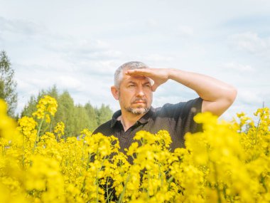  Düşünceli bir ifadeye sahip bir adam çiçek açan bir kolza tohumu tarlasında duruyor, bir eli başının arkasında, yumuşak, parçalı bulutlu bir gökyüzünün altında dinleniyor..
