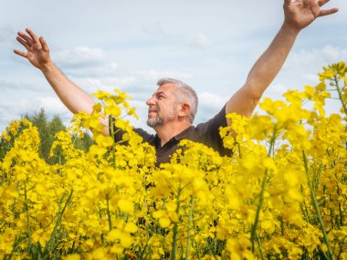 Gülümseyen bir adam, bulutlu bir gökyüzünün altında parlak sarı bir kolza tohumu tarlasında dururken mutlu bir şekilde ellerini kaldırır..