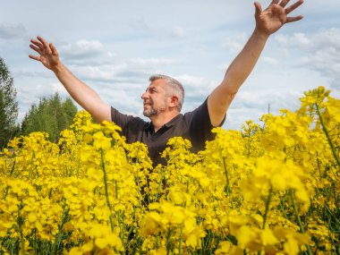 Gülümseyen bir adam, bulutlu bir gökyüzünün altında parlak sarı bir kolza tohumu tarlasında dururken mutlu bir şekilde ellerini kaldırır..