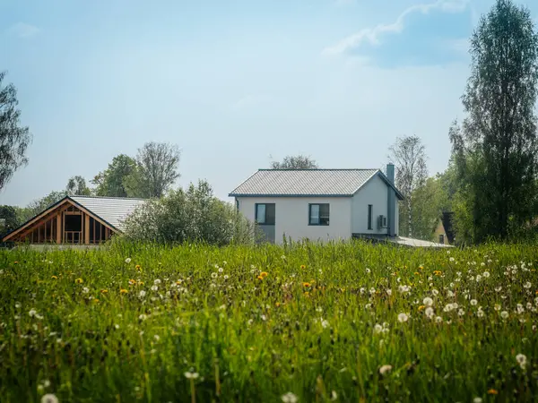 Karahindiba tohumu başları ve sarı çiçeklerle dolu parlak yeşil bir çayır biri hala yapım aşamasında olan iki modern eve doğru uzanır, açık mavi gökyüzüne karşı..