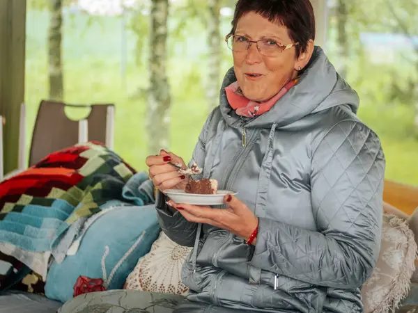 Açık gri örtülü ceketli ve gözlüklü bir kadın kapalı bir bankta oturuyor, elinde çikolatalı pasta tabağı, yastıklarla ve yumuşak ışıklandırmalarla.