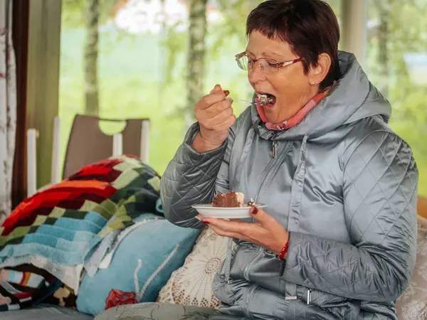 Açık gri örtülü ceketli ve gözlüklü bir kadın kapalı bir bankta oturuyor, elinde çikolatalı pasta tabağı, yastıklarla ve yumuşak ışıklandırmalarla.