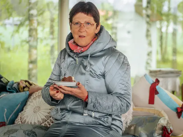 Açık gri örtülü ceketli ve gözlüklü bir kadın kapalı bir bankta oturuyor, elinde çikolatalı pasta tabağı, yastıklarla ve yumuşak ışıklandırmalarla.