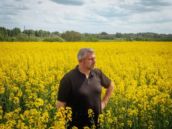 Koyu renk gömlekli düşünceli bir adam bulutlu bir bahar gökyüzünün altında çiçek açan sarı çiçeklerle çevrili geniş bir kolza tohumu tarlasında duruyor..