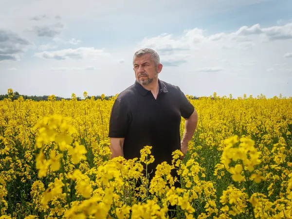 Koyu renk gömlekli düşünceli bir adam bulutlu bir bahar gökyüzünün altında çiçek açan sarı çiçeklerle çevrili geniş bir kolza tohumu tarlasında duruyor..