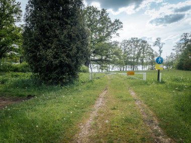 Çimenli bir açıklıktan geçen toprak bir yol, bulutlu bir gökyüzünün altında ağaçlar ve karahindibalarla çevrili, girişi kısıtlayan işaretlerle dolu, kapalı beyaz bir kapıda son bulur..