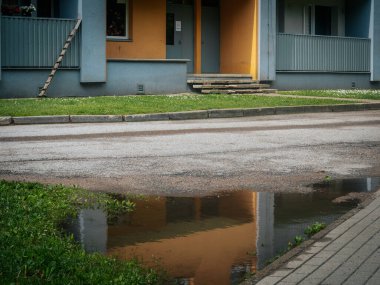 Merdivenleri ve balkonları olan bir konut binasının girişi, sokakta çimen ve kaldırımla çevrili bir su birikintisine yansıyor. Bu sahne yeni yağan yağmuru anımsatıyor..