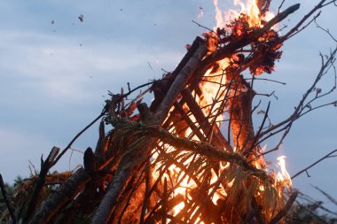 Kütükler ve dallarla inşa edilmiş büyük bir şenlik ateşi alacakaranlıkta parlak bir şekilde yanar. Dekoratif bir çelenk veya bitki demeti yapıda kısmen görülebilir.