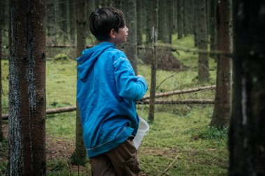 Mavi kapüşonlu genç bir çocuk, uzun orman ağaçları ve yemyeşil ağaçlarla çevrili plastik bir kova tutarken yosun kaplı bir kütüğün üzerinde tek bacağının üzerinde dengede duruyor. Valmiera, Letonya - 25.06.2025