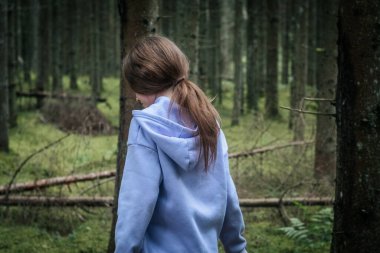 Genç bir kız, açık mavi kapüşonlu ve desenli pantolonlu, uzun ağaçlar ve yumuşak yeşilliklerle çevrili plastik bir konteynır ile yosun kaplı bir ormanda yürüyor..