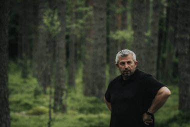 Gri saçlı ve sakallı olgun bir adam sık bir ormanda elleri kalçasında duruyor, siyah bir gömlek giyiyor ve ciddi bir ifadeyle hafifçe yan tarafa bakıyor..