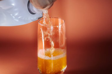 Turuncu köpüklü bir içeceğin yakın çekimi beyaz bir torbadan temiz bir bardağa dökülüyor, sıcak turuncu arka planda..