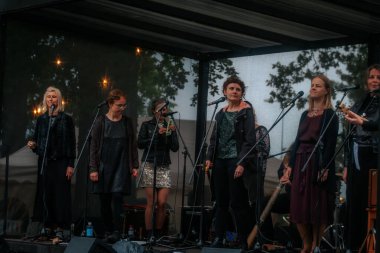 Sahnede bir grup kadın, açık hava müzik etkinliği sırasında, koyu renk giysiler içinde, bir tepe örtüsü altında mikrofon ve enstrümanlarla sahne alıyor..
