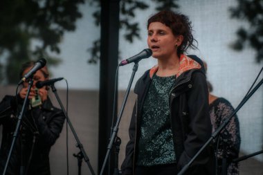 Sahnede bir grup kadın, açık hava müzik etkinliği sırasında, koyu renk giysiler içinde, bir tepe örtüsü altında mikrofon ve enstrümanlarla sahne alıyor..