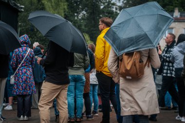 Bir grup insan yağmurlu bir havada açık havada şemsiye ve yağmurluğun altında birbirlerine yakın dururlar..