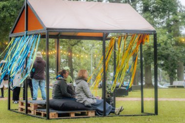 Mavi ve sarı kurdeleli küçük bir açık hava pavyonu yeşil park ortamındaki bir etkinlik sırasında insanları minderlere oturttu..