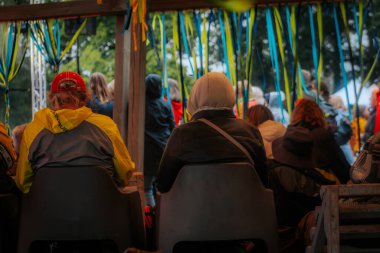 İnsanlar, mavi ve sarı şeritlerle süslenmiş bir gölgeliğin altında, bulutlu bir günde açık hava etkinliğini izleyen sandalyelere otururlar..