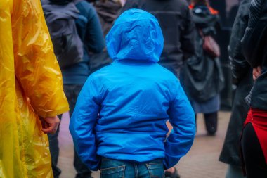 Parlak mavi kapüşonlu küçük bir çocuk kalabalığın içinde öne bakıyor. Bu da sarı pançolu birini de kapsıyor..