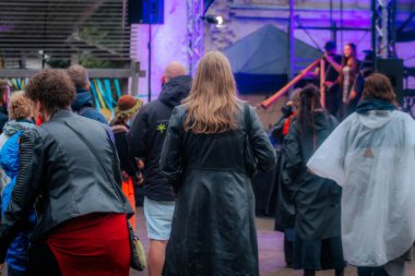 Yağmurluk ve ceket giyen çeşitli insanlar bir açık hava etkinliğinde toplanıyor; bazıları telefonlarını kontrol ediyor ve şemsiye taşıyor. Arkaplanda renkli kurdeleler asılı..