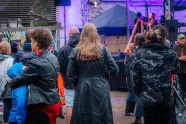 Yağmurluk ve ceket giyen çeşitli insanlar bir açık hava etkinliğinde toplanıyor; bazıları telefonlarını kontrol ediyor ve şemsiye taşıyor. Arkaplanda renkli kurdeleler asılı..